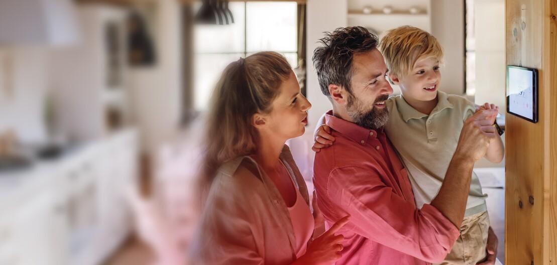 Ein Frau und ein Mann mit einem Kind auf dem Arm stellen die Temperatur an einem Smart Home-Regler ein Ein Frau und ein Mann mit einem Kind auf dem Arm stellen die Temperatur an einem Smart Home-Regler ein