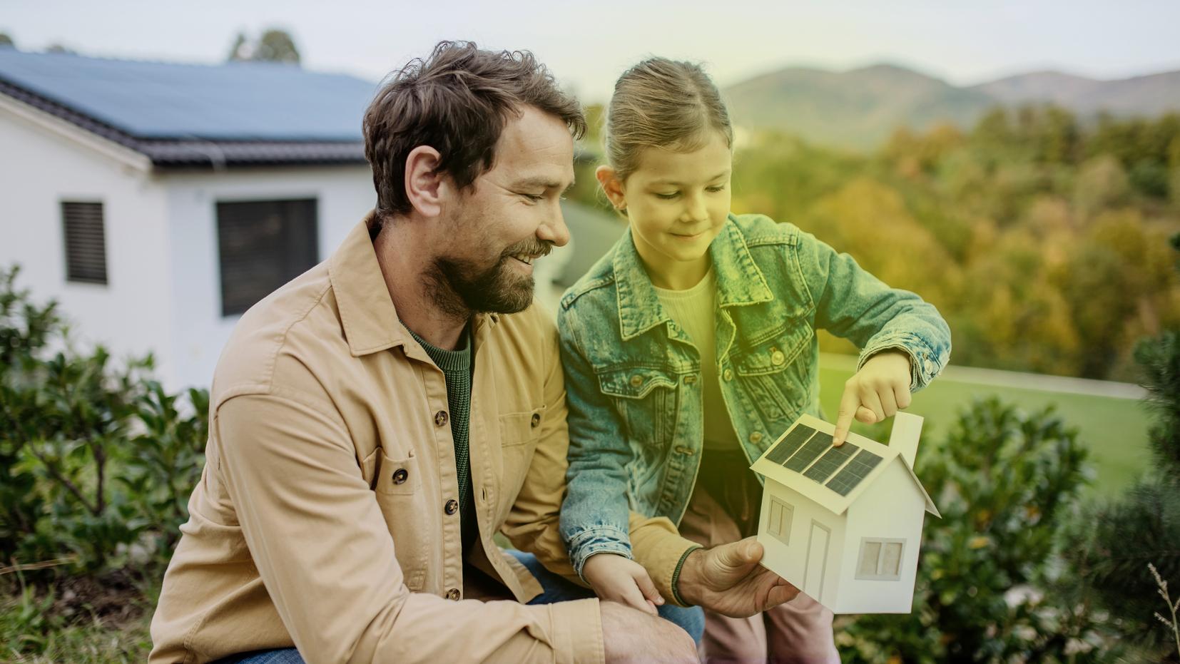 Ein Mann und ein kleines Mädchen stehen im Garten und schauen sich ein Modellhaus mit Photovoltaik auf dem Dach an. Ein Mann und ein kleines Mädchen stehen im Garten und schauen sich ein Modellhaus mit Photovoltaik auf dem Dach an.