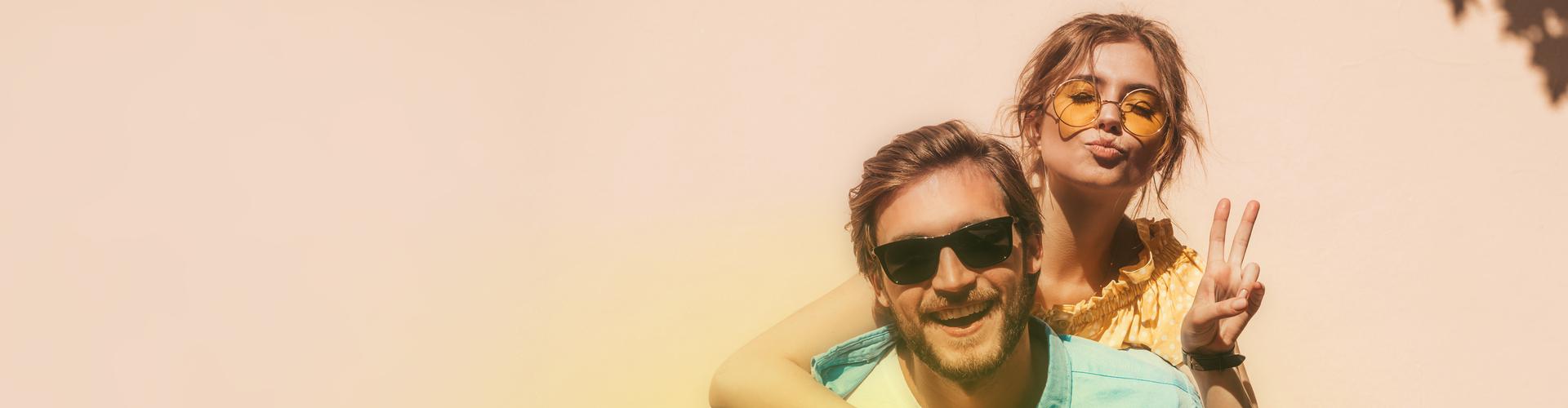 Ein junger Mann mit Sonnenbrille l&auml;chelt fr&ouml;hlich in die Kamera und tr&auml;gt eine Frau Huckepack
