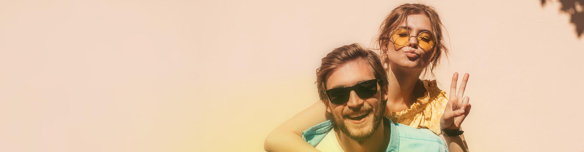 Ein junger Mann mit Sonnenbrille l&auml;chelt fr&ouml;hlich in die Kamera und tr&auml;gt eine Frau Huckepack