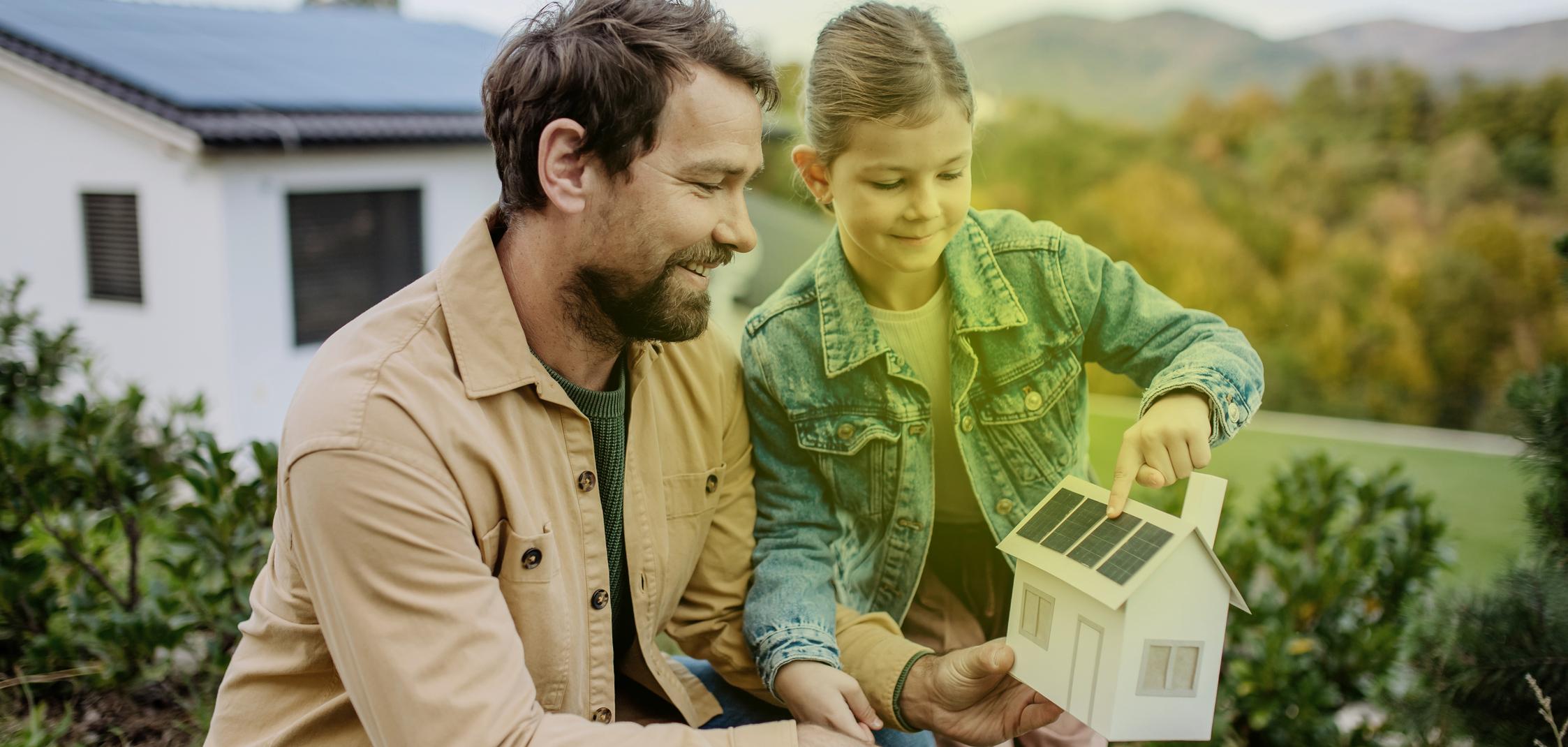 Ein Mann und ein kleines Mädchen stehen im Garten und schauen sich ein Modellhaus mit Photovoltaik auf dem Dach an.