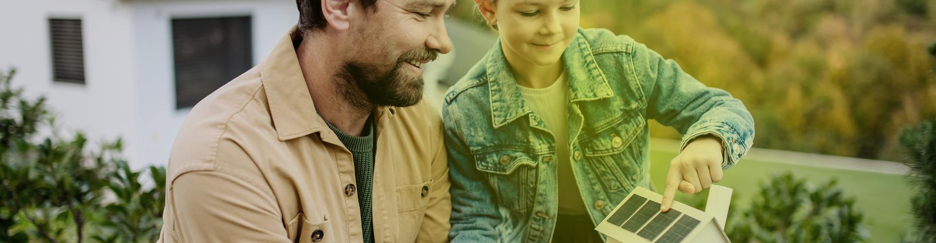 Ein Mann und ein kleines Mädchen stehen im Garten und schauen sich ein Modellhaus mit Photovoltaik auf dem Dach an.