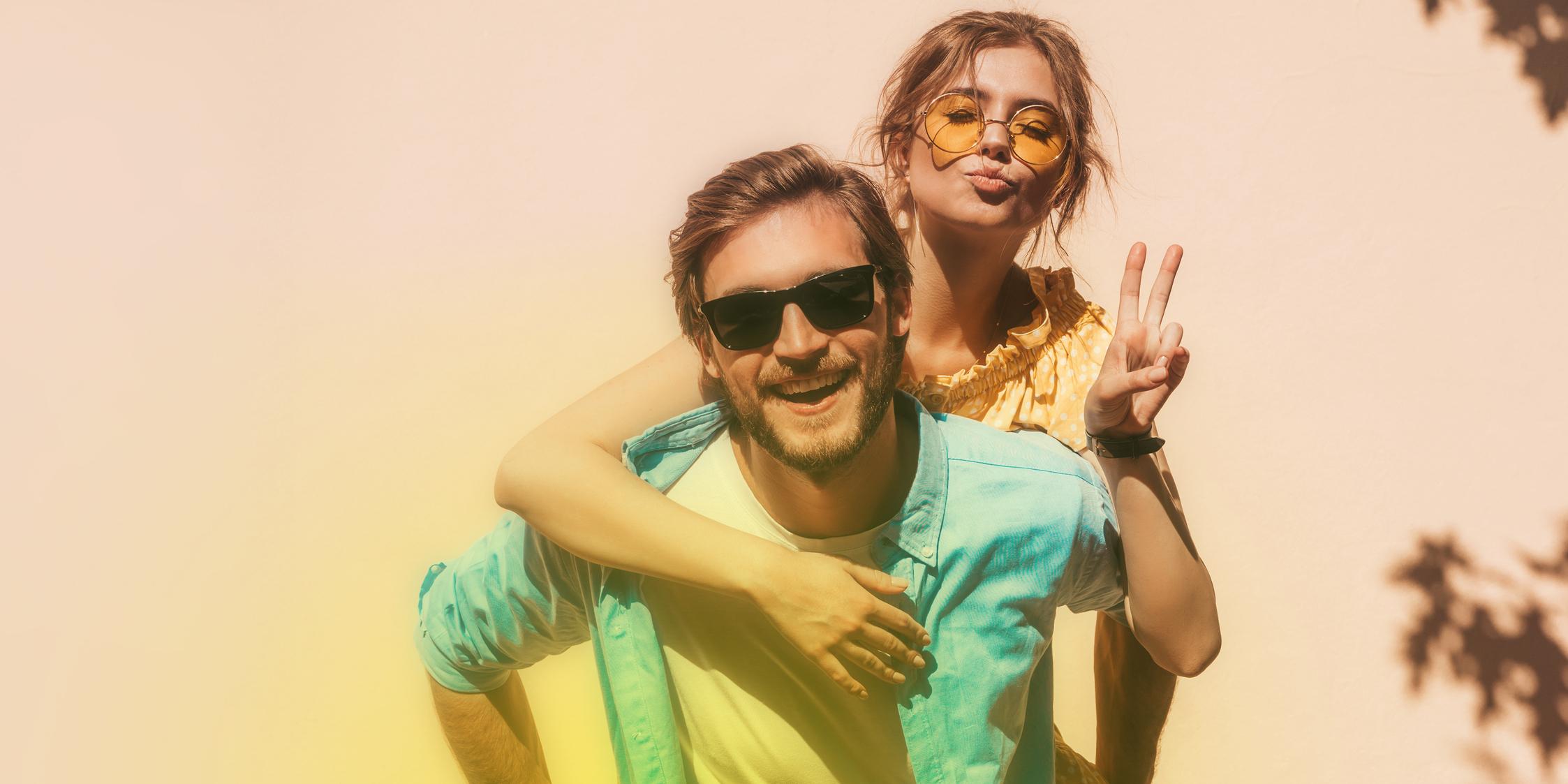 Ein junger Mann mit Sonnenbrille lächelt fröhlich in die Kamera und trägt eine Frau Huckepack