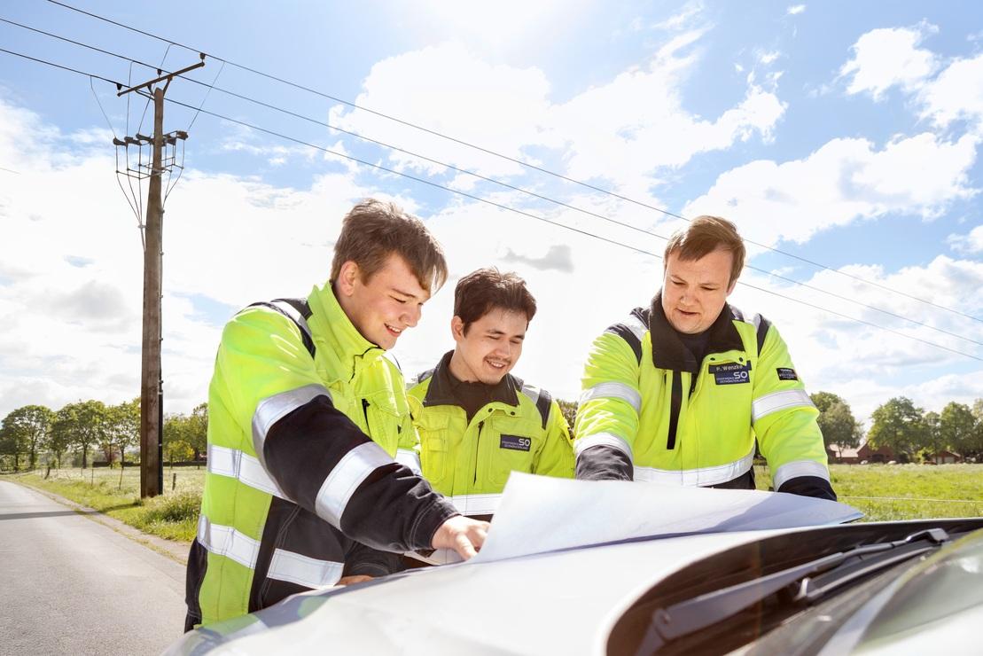 Drei Mitarbeiter der Stadtwerke Ostmünsterland besprechen Pläne auf einer Motorhaube im ländlichen Außenbereich neben einer Straße und Stromleitung Drei Mitarbeiter der Stadtwerke Ostmünsterland besprechen Pläne auf einer Motorhaube im ländlichen Außenbereich neben einer Straße und Stromleitung